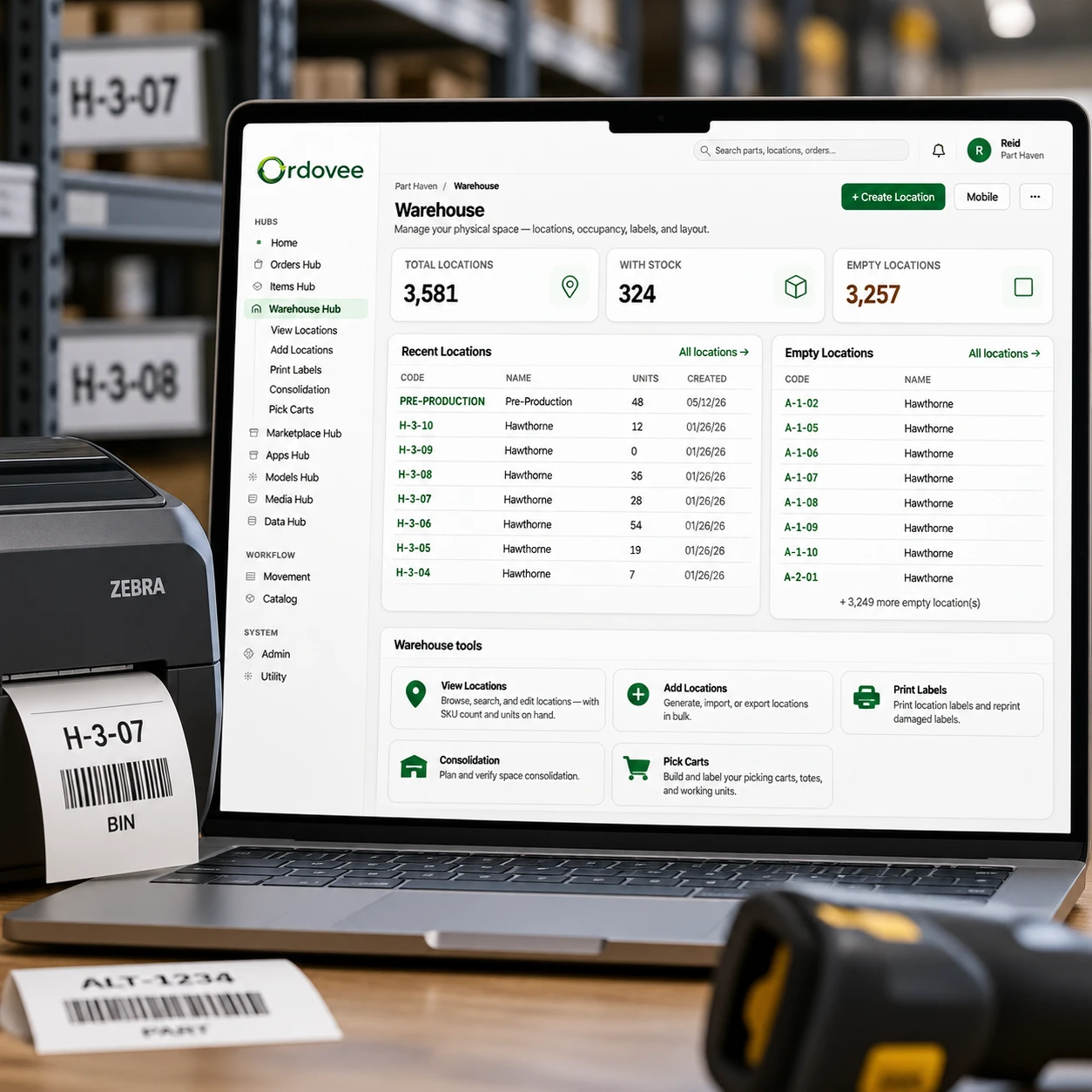 The Ordovee Warehouse hub on a laptop — 3,581 locations, 324 with stock, recent and empty locations listed by code, and the warehouse tools — beside a Zebra label printer feeding out an H-3-07 bin label, a printed ALT-1234 part label, a barcode scanner, and shelving with matching location labels.