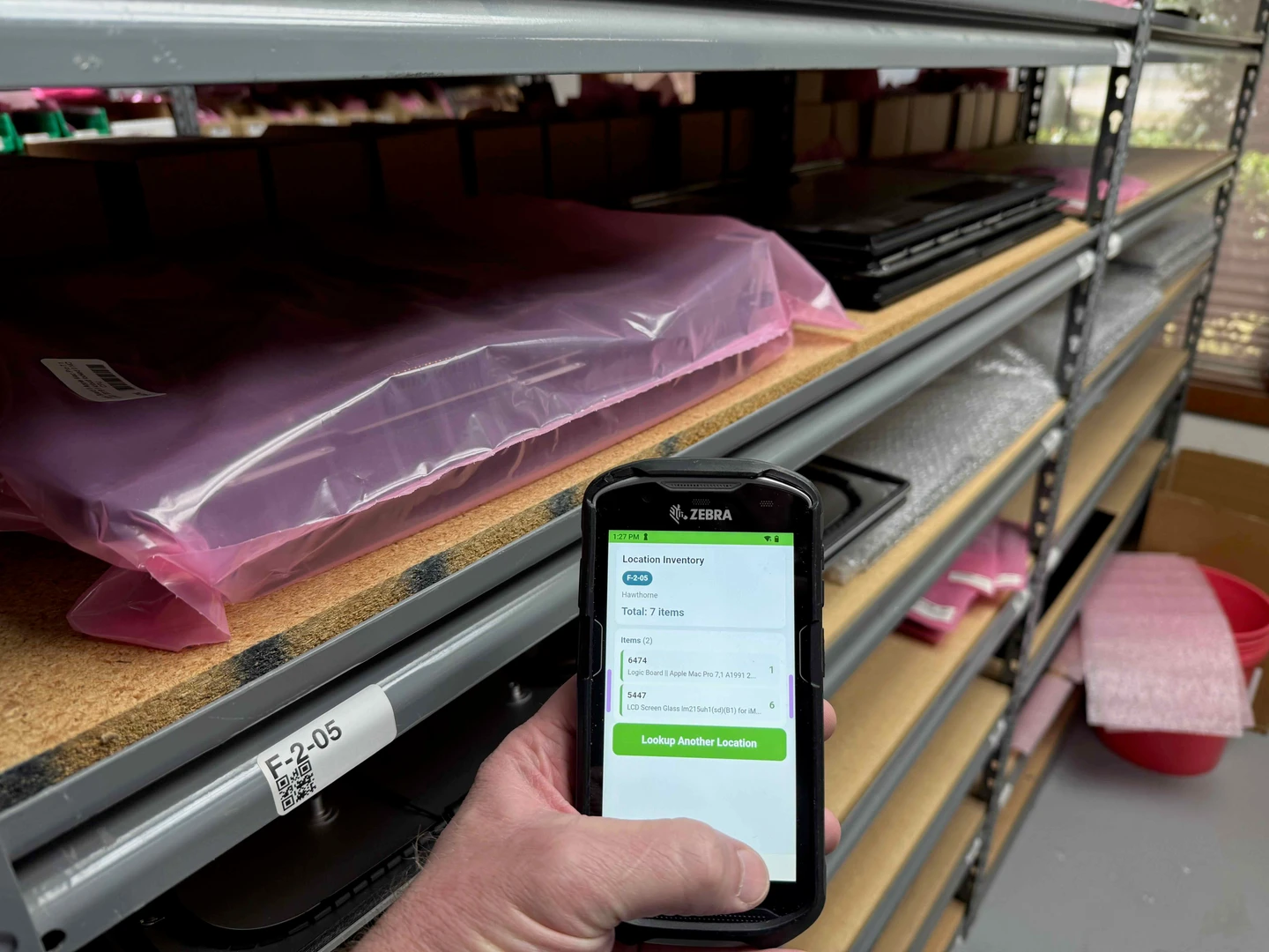 A Zebra TC-series handheld held in a parts warehouse aisle, showing Ordovee's Location Inventory screen for bin F-2-05 with seven items, beside the scanned F-2-05 QR label on the shelf beam