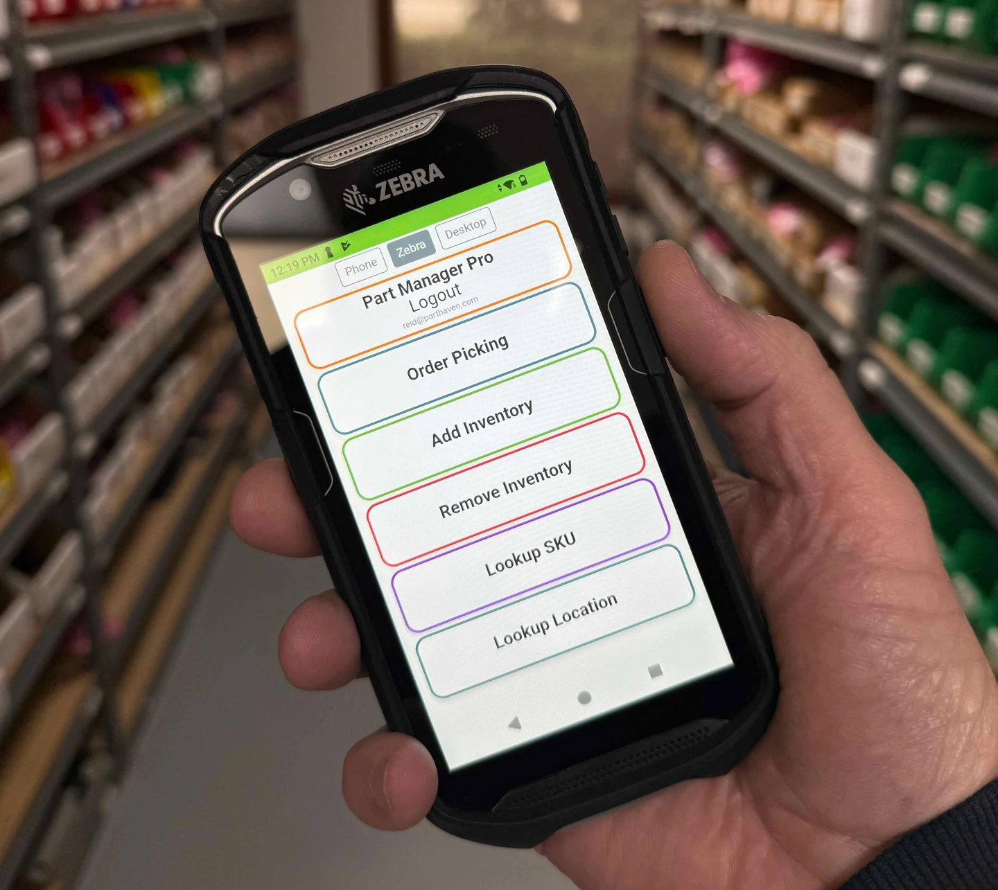 A Zebra TC-series handheld showing the Ordovee home screen, held in a parts warehouse aisle